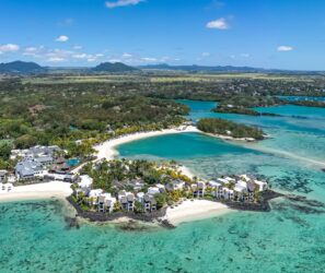 Letecký pohled na&nbsp;tropický plážový resort s&nbsp;bílým pískem, tyrkysovou vodou a&nbsp;vilami obklopenými palmami a&nbsp;bujnou zelení, v&nbsp;dálce jsou vidět hory pod modrou oblohou.