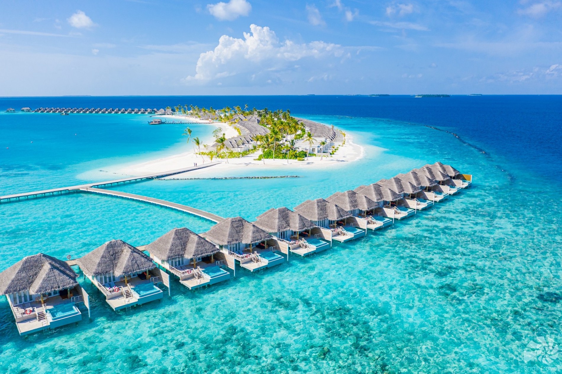 Letecký pohled na&nbsp;bungalovy nad vodou na&nbsp;tyrkysové vodě, propojené dřevěným chodníkem s&nbsp;bílým písečným ostrovem s&nbsp;palmami, obklopené tmavě modrým oceánem pod jasně modrou oblohou.