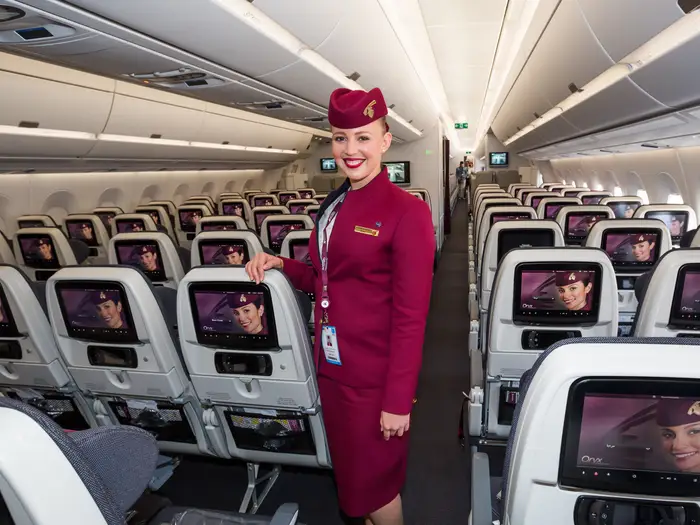 Usměvavá letuška ve&nbsp;vínové uniformě a&nbsp;klobouku stojí v&nbsp;uličce kabiny letadla, obklopená řadami sedadel pro&nbsp;cestující s&nbsp;obrazovkami osobní zábavy.