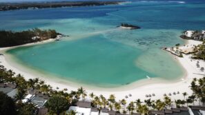 Letecký pohled na&nbsp;tropickou pláž s&nbsp;bílým pískem, tyrkysovou vodou, palmami a&nbsp;slaměnými slunečníky, lemovanou bujnou zelení, malými ostrůvky a&nbsp;několika budovami podél pobřeží.