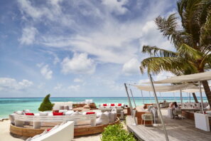 Venkovní plážové posezení s&nbsp;bílým kruhovým posezením, červenými polštáři, palmami a&nbsp;výhledem na&nbsp;tyrkysovou vodu oceánu pod částečně zataženou oblohou. Někteří lidé sedí a&nbsp;odpočívají u&nbsp;baru pod širým nebem.