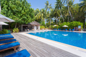 Tropický venkovní bazén s&nbsp;modrými lehátky, obklopený palmami a&nbsp;zelení. Na&nbsp;vzdálenějším konci je zastíněná barová zóna a&nbsp;stoly s&nbsp;bílými slunečníky a&nbsp;průzračně modrou oblohou nad nimi.