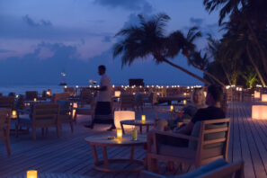 Za soumraku sedí pár u&nbsp;stolu se svíčkami na&nbsp;dřevěné terase u&nbsp;oceánu, obklopen palmami a&nbsp;svítícími lucernami, zatímco kolem prochází číšník a&nbsp;ostatní stoly zůstávají prázdné.