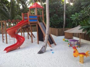 Dětské hřiště s&nbsp;červenou skluzavkou, prolézačkami a&nbsp;zastíněným pískovištěm na&nbsp;bílém písku, obklopené palmami a&nbsp;zelení. Je zde žluté pružinové houpadlo a&nbsp;malý stolek se židlemi.