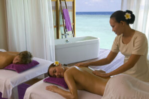 V klidné místnosti s&nbsp;výhledem na&nbsp;oceán masíruje lázeňský terapeut ženu zad, zatímco další osoba relaxuje na&nbsp;vedlejším masážním stole. Klidnou atmosféru dotváří vana a&nbsp;bílé závěsy.