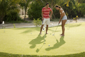 Muž a&nbsp;žena hrají minigolf na&nbsp;hřišti obklopeném palmami. Muž v&nbsp;růžovém pruhovaném tričku a&nbsp;šortkách sleduje ženu v&nbsp;bílém topu a&nbsp;šortkách, která se připravuje k&nbsp;puttování na&nbsp;slunném hřišti naboso.