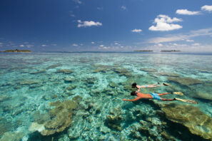 Dva lidé šnorchlují nad průzračným, mělkým korálovým útesem pod jasně modrou oblohou s&nbsp;roztroušenými mraky, obklopeni tyrkysovou vodou a&nbsp;vzdálenými ostrůvky.