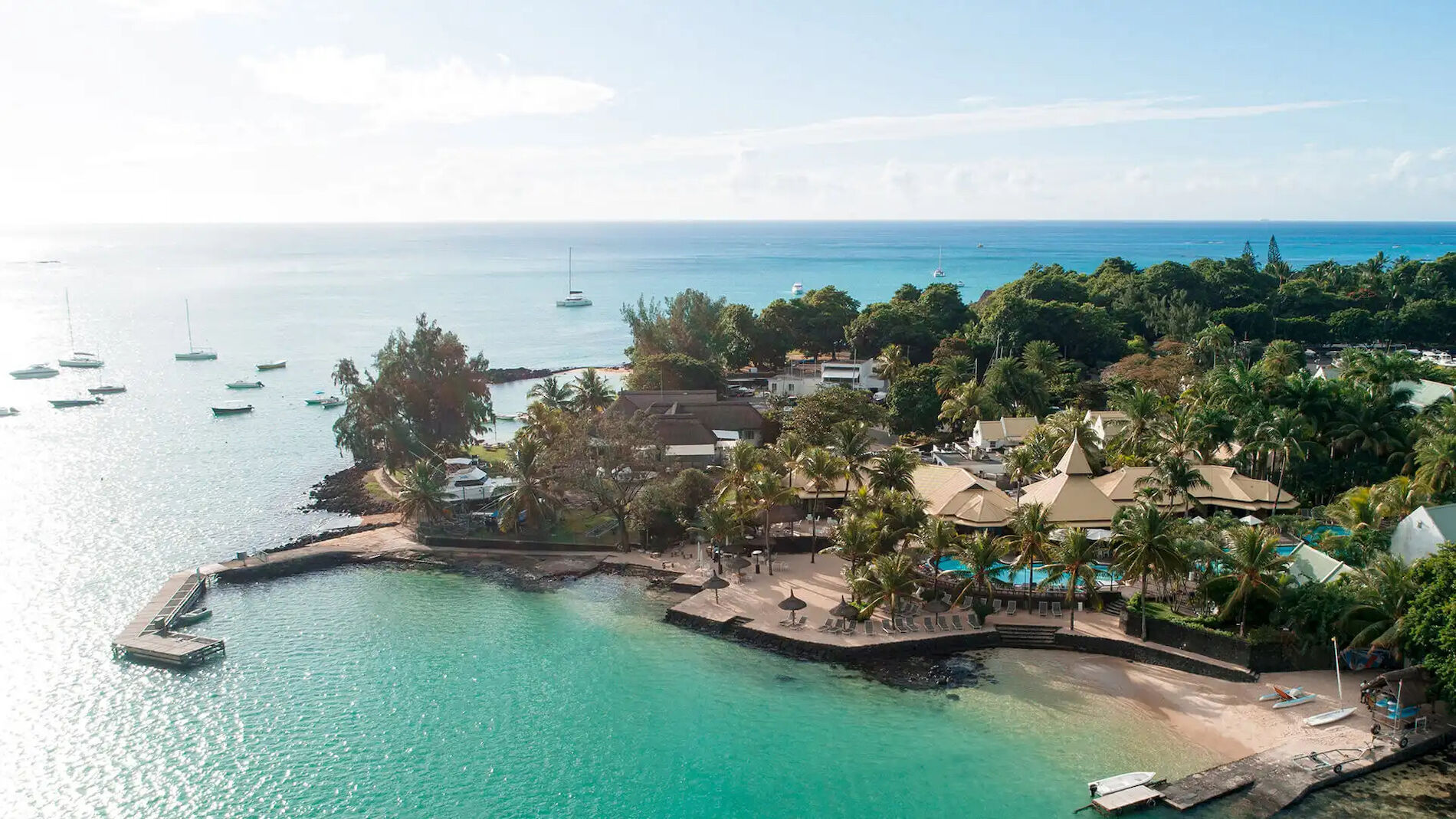 Letecký pohled na&nbsp;tropický plážový resort s&nbsp;tyrkysovou vodou, palmami, lehátky, bazény a&nbsp;loděmi kotvícími na&nbsp;pobřeží pod jasnou slunečnou oblohou.