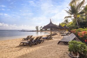 Písečná pláž s&nbsp;řadami prázdných lehátek pod slaměnými slunečníky, palmami a&nbsp;barevnými květinami po&nbsp;stranách. V&nbsp;pozadí klidná voda oceánu a&nbsp;částečně zatažená obloha.