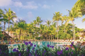 Tropický venkovní bazén obklopený palmami a&nbsp;lehátky s&nbsp;fialovými květy v&nbsp;popředí pod jasně modrou oblohou.