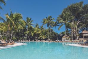 Velký venkovní bazén obklopený palmami a&nbsp;lehátky pod jasně modrou oblohou vytváří tropickou a&nbsp;relaxační atmosféru letoviska.