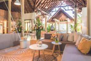 Světlý, otevřený salonek s&nbsp;béžovými pohovkami, kulatými bílými stoly, svěží květinovou výzdobou a&nbsp;dřevěnými stropy s&nbsp;vysokými trámy. Dovnitř proudí sluneční světlo, které zvýrazňuje tropické rostliny a&nbsp;osobu v&nbsp;pozadí u&nbsp;vchodu.