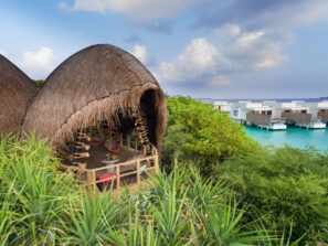 Ze slaměné chýše ve&nbsp;tvaru kopule pod širým nebem je výhled na&nbsp;bujnou zeleň a&nbsp;tyrkysovou vodu, v&nbsp;pozadí se pod částečně zataženou oblohou tyčí moderní bílé vily nad vodou.