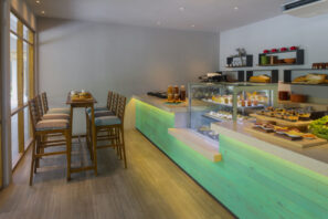 Moderní kavárna s&nbsp;dřevěnou podlahou, vysokými stoly a&nbsp;židlemi, skleněnou vitrínou plnou zákusků a&nbsp;dezertů a&nbsp;policemi s&nbsp;nádobím a&nbsp;dekoracemi. Pod pultem svítí nazelenalá záře.