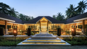 Moderní budova tropického resortu s&nbsp;vrcholovou střechou, jídelnami pod širým nebem a&nbsp;světly svítícími za&nbsp;soumraku, obklopená palmami a&nbsp;zapálenými pochodněmi lemujícími vstupní cestu.