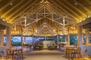 Prostorná moderní restaurace s&nbsp;vysokým klenutým dřevěným stropem, bílými trámy, teplým osvětlením, dřevěnými stoly a&nbsp;židlemi a&nbsp;otevřenými stěnami, které odhalují venkovní svěží zeleň.