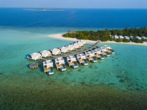 Letecký pohled na&nbsp;vily nad vodou a&nbsp;bungalovy na&nbsp;pláži v&nbsp;tropickém letovisku, obklopeném průzračným tyrkysovým mořem a&nbsp;bujnou zelení, s&nbsp;dřevěným chodníkem spojujícím vily s&nbsp;ostrovem.