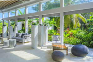 Světlý, moderní salonek s&nbsp;bílou pískovou podlahou, minimalistickým nábytkem, vysokými mramorovými sloupy a&nbsp;velkými okny s&nbsp;výhledem na&nbsp;svěží tropickou zeleň a&nbsp;palmy. Prostor naplňuje přirozené světlo.