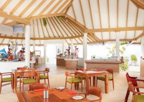 Světlá, vzdušná restaurace s&nbsp;vysokým trámovým stropem, dřevěným nábytkem se zelenými polštáři, pískovou podlahou a&nbsp;bufetovými stoly v&nbsp;pozadí; velká okna propouštějí přirozené světlo.