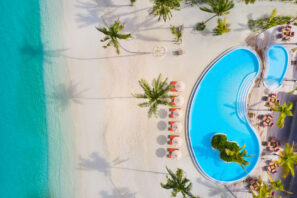 Letecký pohled na&nbsp;tropický plážový resort s&nbsp;tyrkysovou vodou, bílým pískem, palmami, červenými slunečníky a&nbsp;zakřiveným bazénem s&nbsp;ostrovem uprostřed.