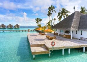 Dřevěná paluba se stoly, židlemi a&nbsp;žlutými slunečníky se rozprostírá nad průzračnou tyrkysovou vodou vedle budov s&nbsp;doškovými střechami a&nbsp;palmami, v&nbsp;pozadí jsou další bungalovy pod vodou a&nbsp;částečně zatažená obloha.