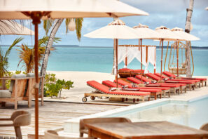Luxusní lounge na&nbsp;pláži s&nbsp;červenými polstrovanými lehátky, bílými slunečníky a&nbsp;dřevěnými kabinami u&nbsp;bazénu s&nbsp;výhledem na&nbsp;tyrkysovou vodu oceánu a&nbsp;písečné pobřeží lemované palmami.