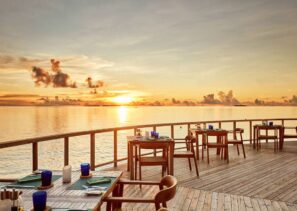 Venkovní terasa restaurace s&nbsp;dřevěnými stoly a&nbsp;židlemi má výhled na&nbsp;klidný oceán při západu slunce. Obloha je oranžová a&nbsp;zlatá s&nbsp;rozptýlenými mraky, což vytváří klidnou a&nbsp;malebnou atmosféru při stolování.