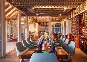 Stylová restaurační jídelna s&nbsp;dlouhým dřevěným stolem pro&nbsp;hosty, obklopená světle modrými židlemi, stojany na&nbsp;víno na&nbsp;stěně a&nbsp;velkými okny, kterými proudí teplé přirozené světlo.