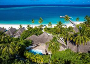 Letecký pohled na&nbsp;tropické plážové letovisko s&nbsp;vilami se slaměnými střechami, palmami, bazénem, bílou písečnou pláží, tyrkysovým oceánem a&nbsp;dlouhým dřevěným molem zasahujícím do&nbsp;moře pod jasně modrou oblohou.