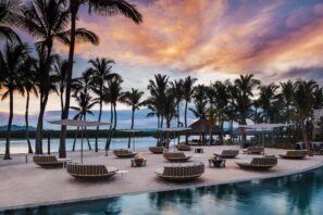 Venkovní lounge u bazénu s kulatým polstrovaným posezením, palmami a ohništěm, s výhledem na vodu při západu slunce, barevnou oblohu a vzdálené hory.