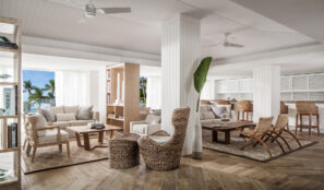 Světlý, vzdušný salonek s proutěným nábytkem, bílými stěnami, velkými okny, tropickými rostlinami a výhledem na venkovní palmy. Prostor má útulné posezení, neutrální tóny a moderní pobřežní design.