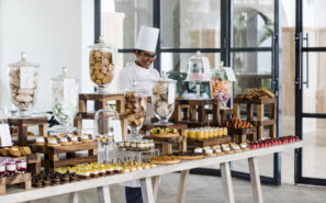 Za stolem plným zákusků, sušenek, pečiva a sladkostí vystavených ve skleněných nádobách a na dřevěných stojanech ve světlé moderní místnosti stojí šéfkuchař.