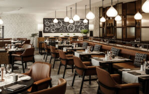 Moderní interiér restaurace s hnědými koženými židlemi, tmavými dřevěnými stoly na stolování, závěsnými světly a dekorativním černobílým nástěnným obrazem v pozadí. Atmosféra je vřelá a příjemná.