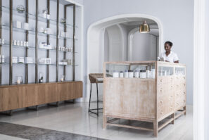 Za světlým dřevěným recepčním pultem v moderním minimalistickém wellness centru s policemi s produkty péče o pleť a jemným neutrálním dekorem stojí žena.