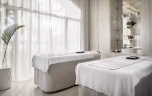 Klidná lázeňská místnost se dvěma masážními stoly pokrytými bílým povlečením, měkkým přirozeným světlem pronikajícím skrz průsvitné závěsy a poličkou s ručníky, lahvemi a rostlinami v pozadí.