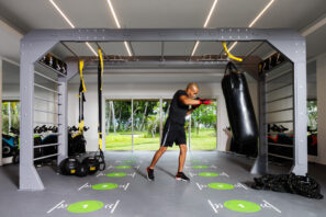 Osoba v černém cvičebním oblečení a červených rukavicích mlátí do těžkého pytle v moderní posilovně s velkými okny, zeleným trávníkem venku a různým fitness vybavením v pozadí.