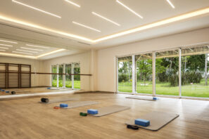 Světlé studio jógy s velkými okny s výhledem do zeleně, čtyřmi jógovými podložkami s bloky a popruhy na dřevěné podlaze a zrcadlovými stěnami odrážejícími prostorný, moderní interiér.