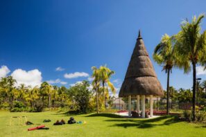 Travnaté hřiště s venkovním fitness vybavením a palmami, na kterém stojí vysoká kuželovitá stavba s doškovou střechou pod jasně modrou oblohou. V pozadí je vidět tenisový kurt.