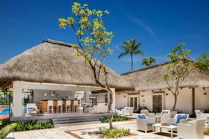 Venkovní tropický lounge prostor s budovami s doškovou střechou, bílým proutěným nábytkem, barovým posezením, zelenými rostlinami a palmami pod jasně modrou oblohou.