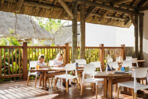 Dvě děti sedí u dřevěného stolu pod doškovou střechou na sluncem ozářené venkovní terase. Kolem nich jsou prázdné bílé židle a stoly, v pozadí bujné zelené rostliny a tropické budovy.