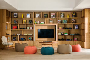 Útulný obývací pokoj s barevnými sedacími vaky s fazolemi, který je obklopen velkou televizí a dřevěnými policemi s knihami, hrami a dekorativními předměty. Vlevo sedí chlupaté bílé křeslo. Místnost působí moderním, přívětivým dojmem.