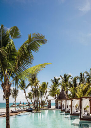 Bazén v tropickém letovisku lemovaný palmami, bílými kabinkami a lehátky s výhledem na jasně modrý oceán pod jasnou oblohou.