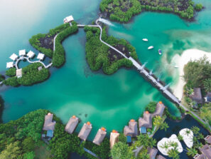 Letecký pohled na bungalovy nad vodou, svěží zelené stromy, klikatící se dřevěný chodník a tyrkysovou lagunu s malými čluny zakotvenými u písečné pláže.