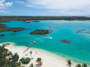 Letecký pohled na tropickou pláž s bílým pískem, tyrkysovou vodou, palmami a malým přístavištěm s čluny. Jeden člun vytváří ve vodě kruhovou stezku, v pozadí jsou svěže zelené ostrovy.