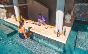 Žena v černých plavkách sedí u baru v bazénu, zatímco barman ve fialové uniformě připravuje nápoje na mramorovém pultu. Na baru jsou rozmístěny láhve, ovoce a dekorativní předměty.