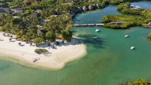 Letecký pohled na tropickou pláž s bílým pískem, tyrkysovou vodou, palmami, malými čluny v laguně, mostem a bujnou zelení kolem pobřeží a blízkých domů.