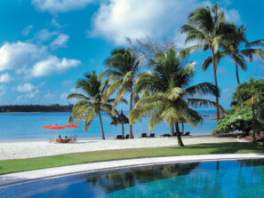 Tropická pláž s palmami, modrým bazénem v popředí, bílým pískem, klidnou vodou oceánu a lidmi odpočívajícími pod oranžovými slunečníky. Obloha je jasně modrá s roztroušenými mraky.