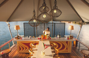 Barman připravuje nápoje ve venkovním baru pod stříškou u vody s moderními závěsnými světly, židlemi a barevnými lahvemi na pultu. Teplé osvětlení a malebný výhled na jezero vytvářejí uvolněnou atmosféru.