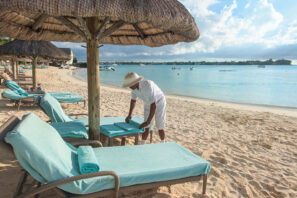 Osoba v&nbsp;bílém oděvu rozkládá tyrkysové ručníky na&nbsp;lehátku pod slaměným slunečníkem na&nbsp;písečné pláži s&nbsp;klidnou modrou vodou a&nbsp;loděmi v&nbsp;pozadí.
