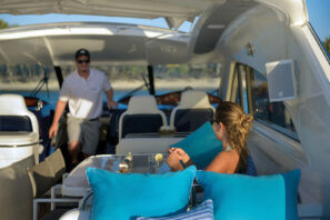 Žena odpočívá na&nbsp;jachtě s&nbsp;modrými polštáři, v&nbsp;ruce drží nápoj a&nbsp;muž v&nbsp;bílé polokošili a&nbsp;čepici stojí u&nbsp;kormidla pod částečně zastíněnou palubou za&nbsp;slunečného dne.